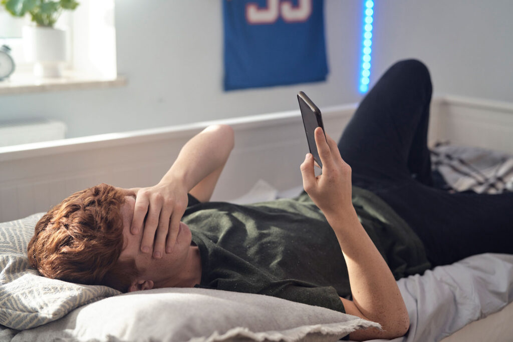 teenage boy covering eyes while browsing phone on bed